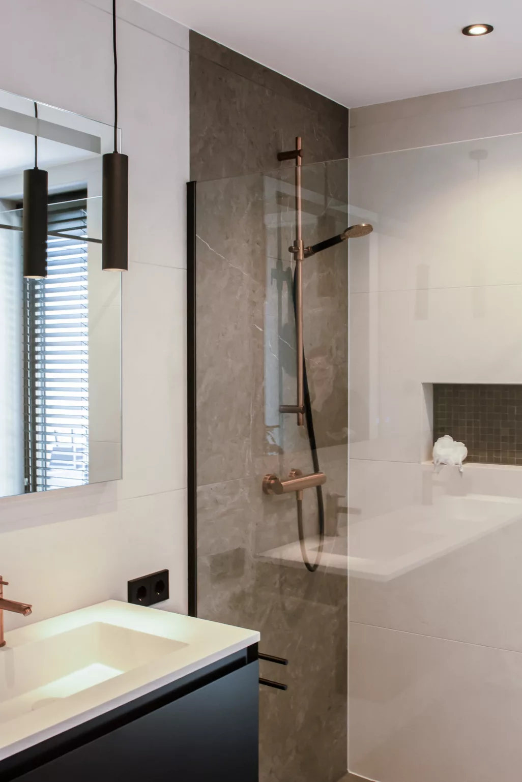 Badkamer met een wastafel met zwart meubel en witte wasbak. De wanden zijn licht. De achterwand van de douche is een aardetint marmer met koperkleurige stang en kraan. 