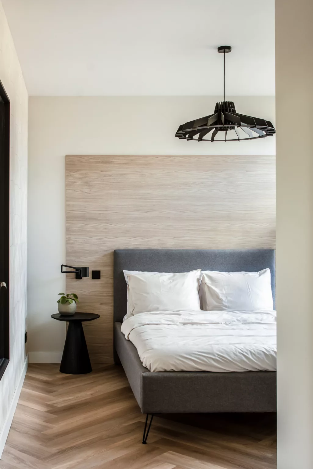 Lichte slaapkamer met visgraatvloer, donkergrijs boxspring en licht beddengoed. Achter het bed is een grote houten wand geplaatst. Het nachtkastje is een klein zwart rond bijzettafeltje. Boven het bed hangt een zwarte draadlamp in organische vorm. 