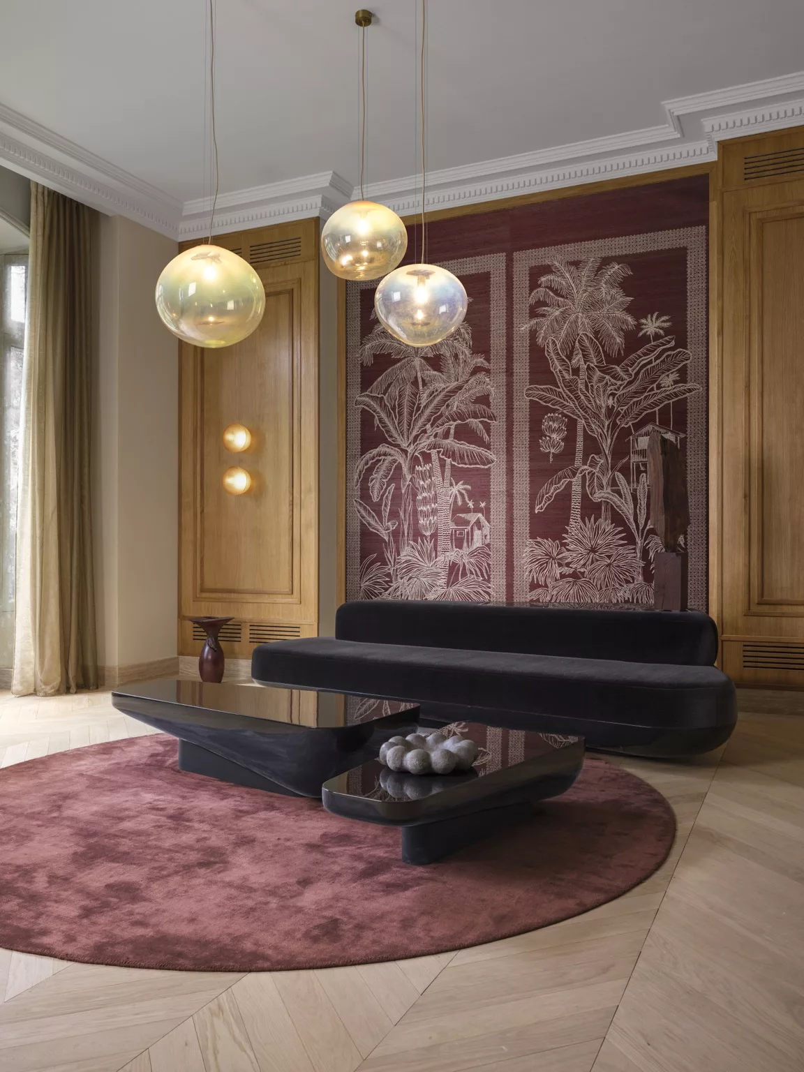 Woonkamer, zwarte velvet bank, rood rond bordeaux vloerkleed, zwart marmer salontafels, achterwand bekleed met zijden behang van bordeauxrode palmbomen