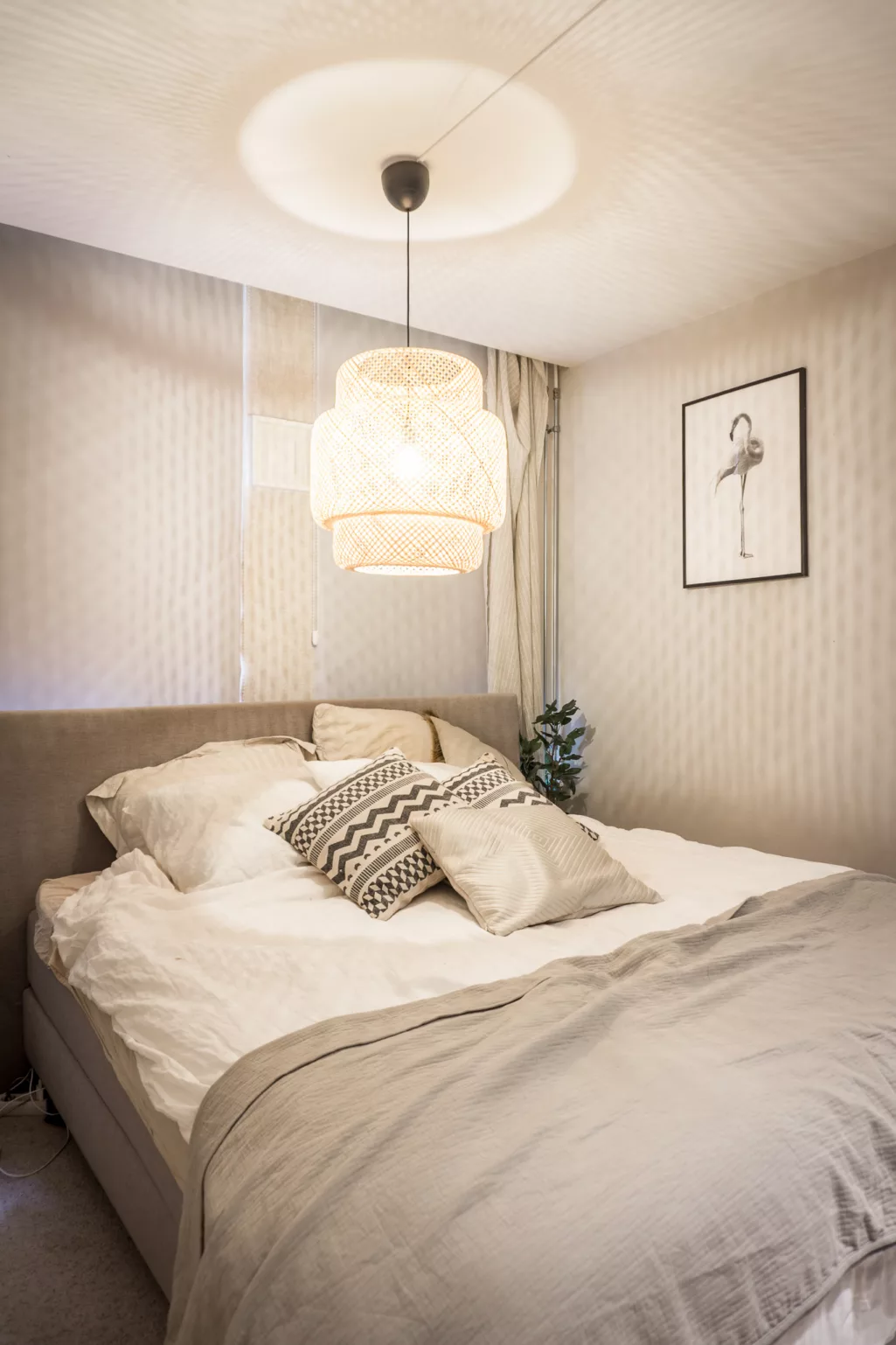 Heel kleine naturellen slaapkamer met een beige behang met licht motief. Een taupe kleurig bed met met hoog headboard. Het bed is opgemaakt met wit bedlinnen, een taupe kleurig sprei en diverse texturen kussens met grafische print en effen beige. Boven het bed hangt een rotan lamp. 