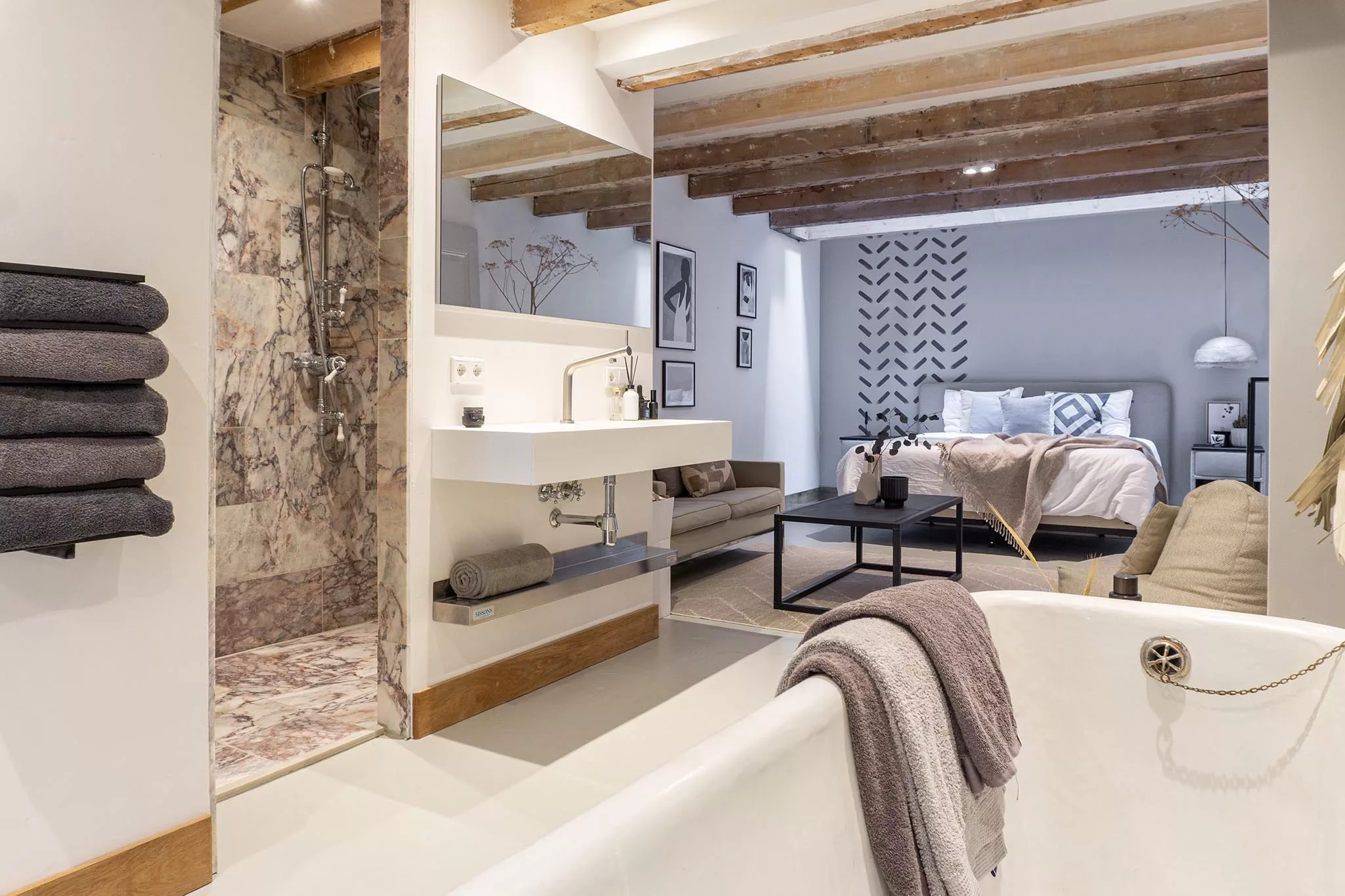 Ensuite slaapkamer met framebed, wasmeubel, inloopdouche en vrijstaande ouderwetse badkuip op pootjes. De kleuren zijn beige en donkerbruin gecombineerd met grijze wanden en houten balken. Het sanitair is wit. 