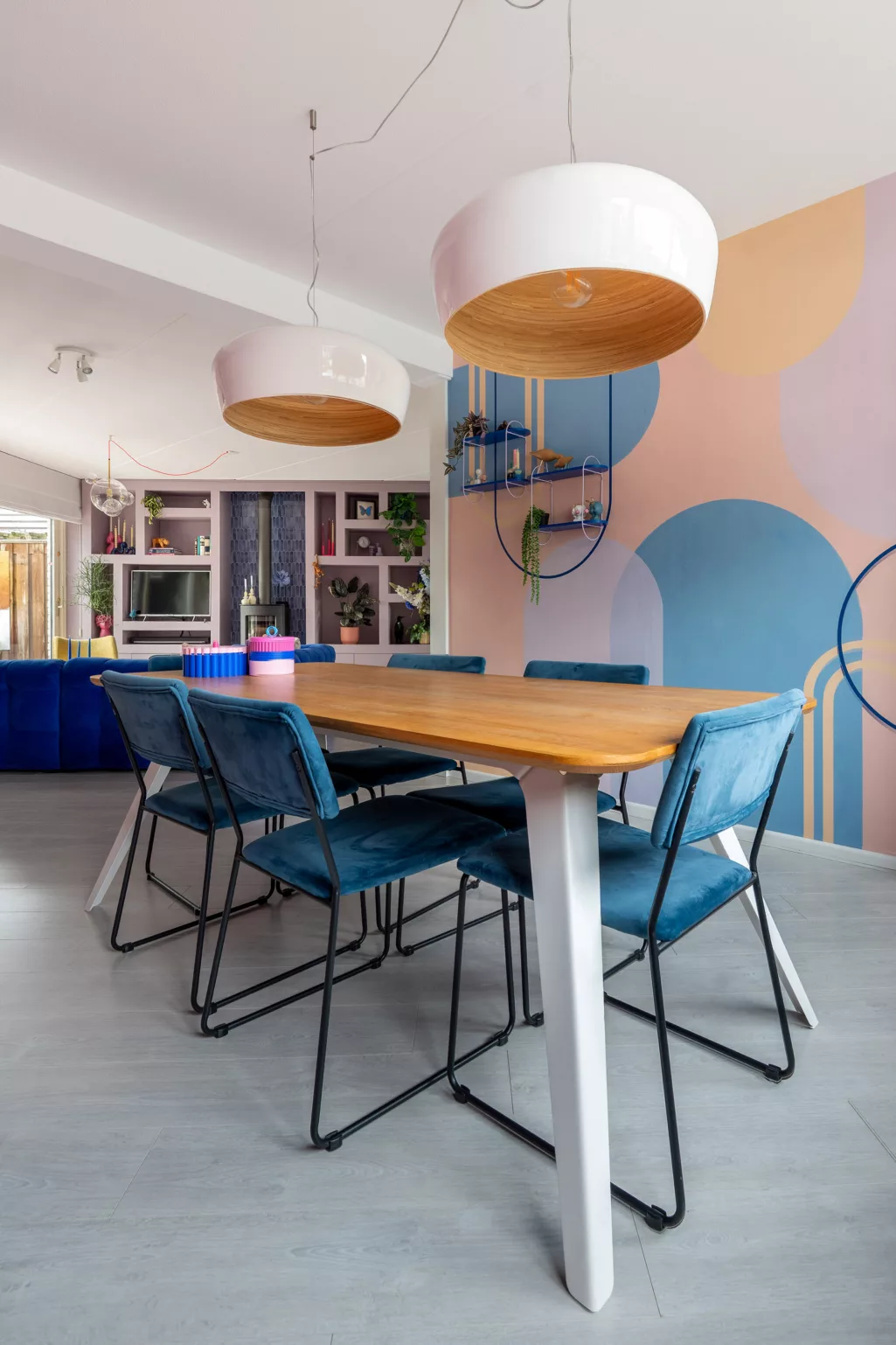 Muur met grafisch behang in bogen, blauw in combinatie met zalmroze en geel, houten eettafel met afgeronde hoeken, eetkamerstoelen in blauw velvet met zwart stalen frame, grote hanglampen wit glanzend met houten binnenkant. 