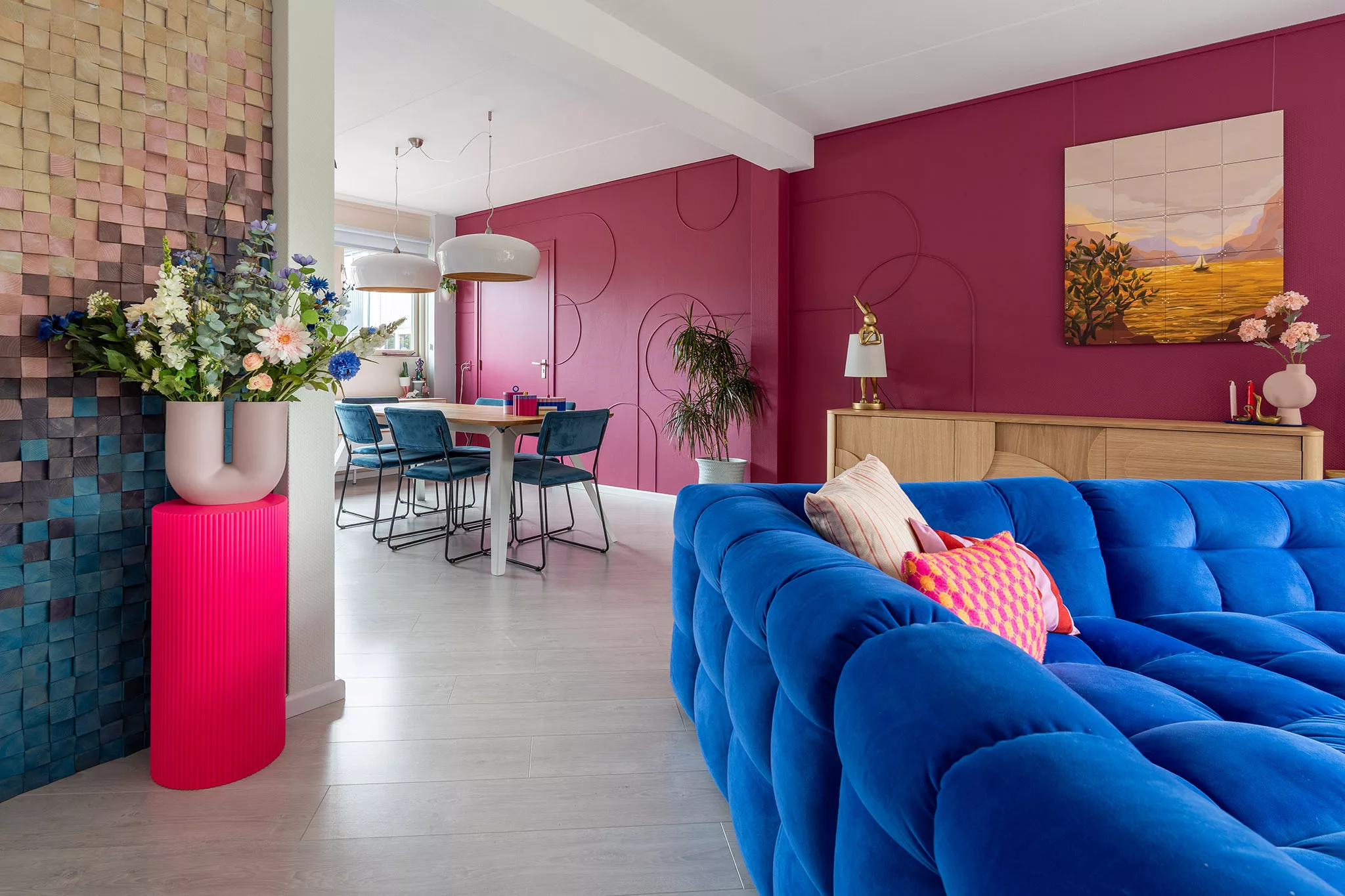 Woonkamer met aubergine kleurige wanden met DIY boogvormen, felblauwe velvetbank, laminaatvloer, neonkleurige sokkel met vaas.