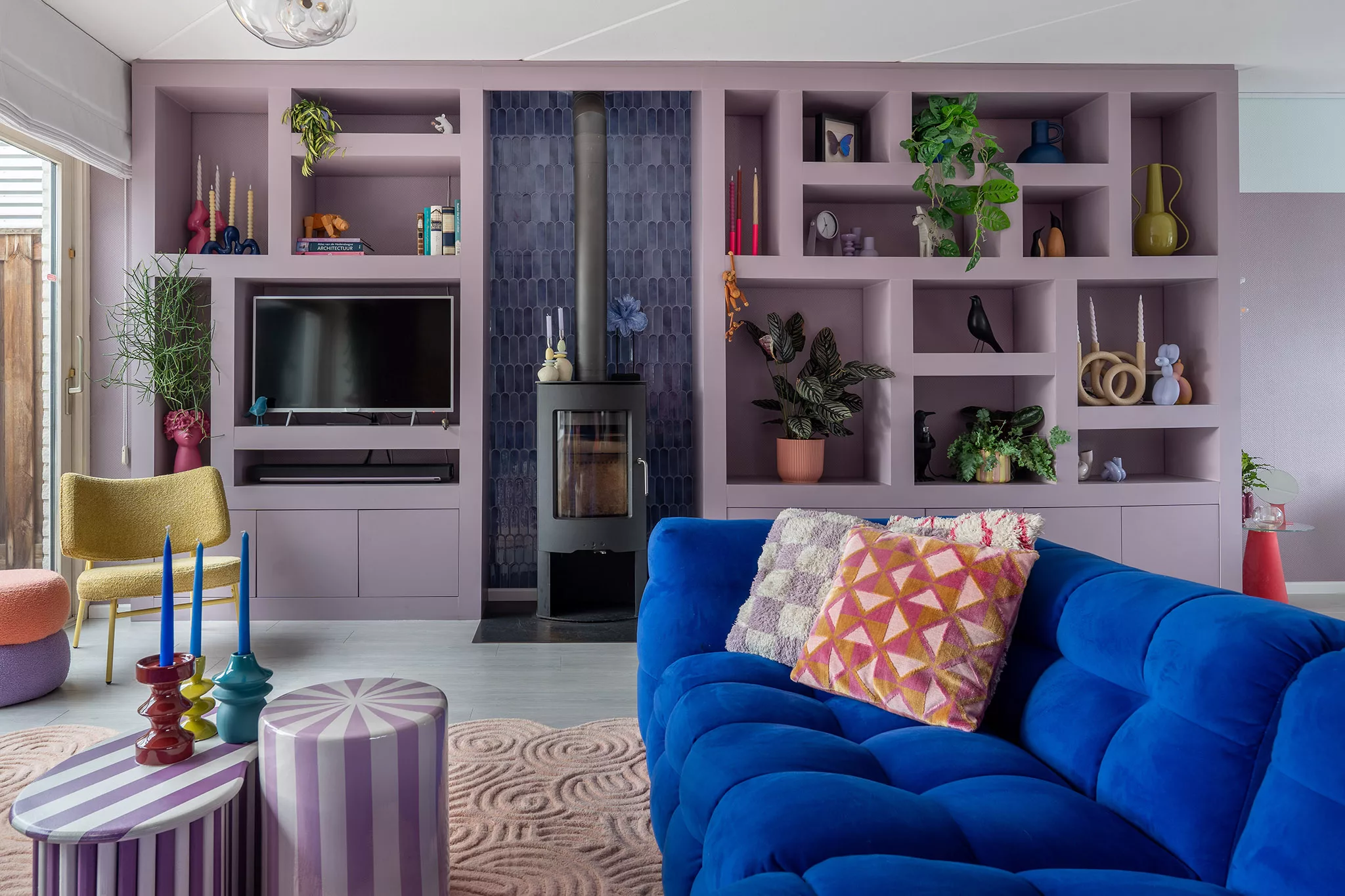Kleurrijk interieur met een lila wandkast, blauwe tegeltjes, kobaltblauwe velvet blikkenbank, lila gestreepte salontafels, muurvlak lila. 