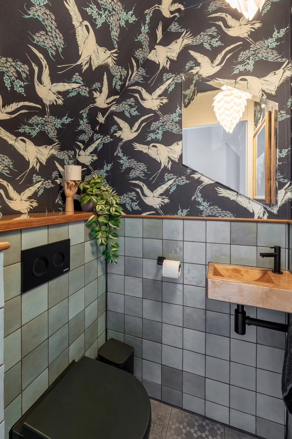 Kleurrijk toilet met tegels in groen- en blauwtinten, donkerblauw behang met vogels, zwart zwevend toilet. 