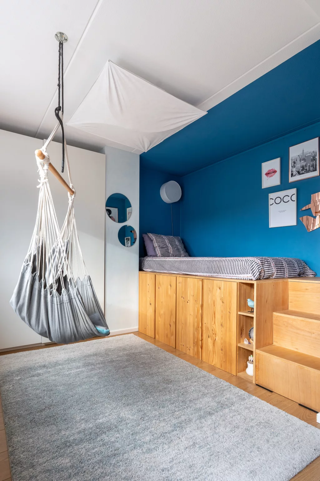Jongenskamer met felblauw op de wand, zelfgemaakt houten bed in nis, hangstoel aan plafond. 