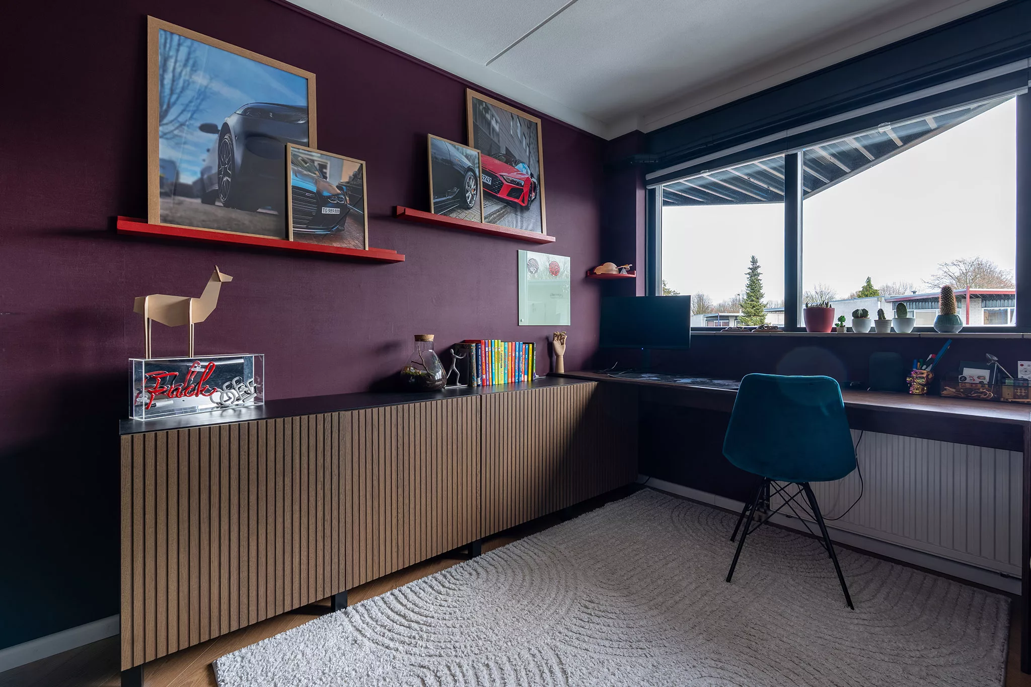 Aubergine kleur wanden, houten latten dressoir, bureau met blauwe velvet bureaustoel, donkerblauw vouwgordijn, wit vloerkleed met bogen. 