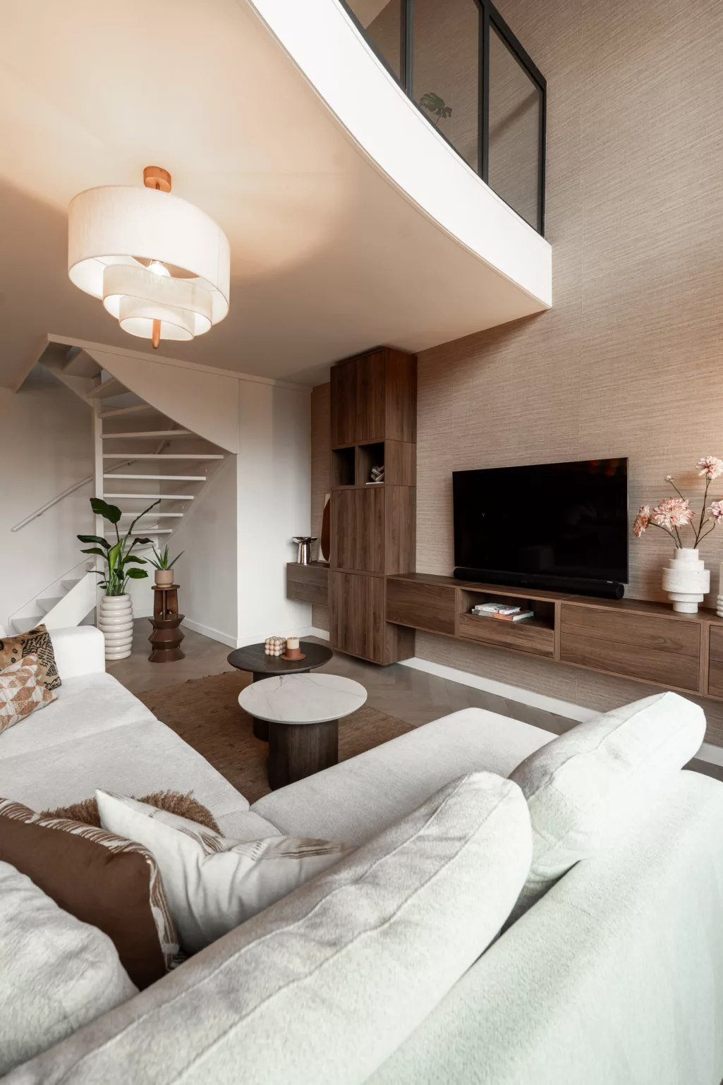 Appartement met hoog plafond, crèmekleurige hoekbank, houten wandmeubel met televisie, open trap wit