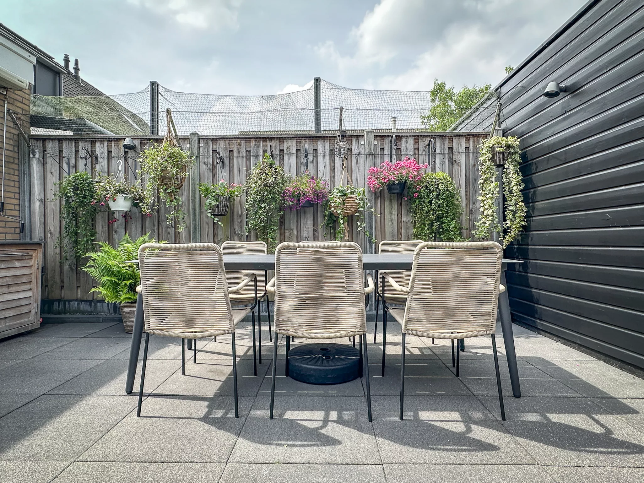rechte tafel met 6 rieten beige stoelen, zwart houten schuur, lichthouten schutting, net voor katten, hangplanten aan schutting