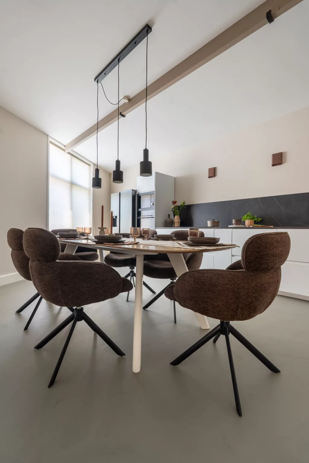 Woonkeuken met eethoek, ovalen tafel visgraat hout, donkerbruine stoffen stoelen met zwart onderstel, beige wanden, balk plafond donker beige, witte keukenkasten, donker natuursteen blad en backsplash, koperen wandlampen