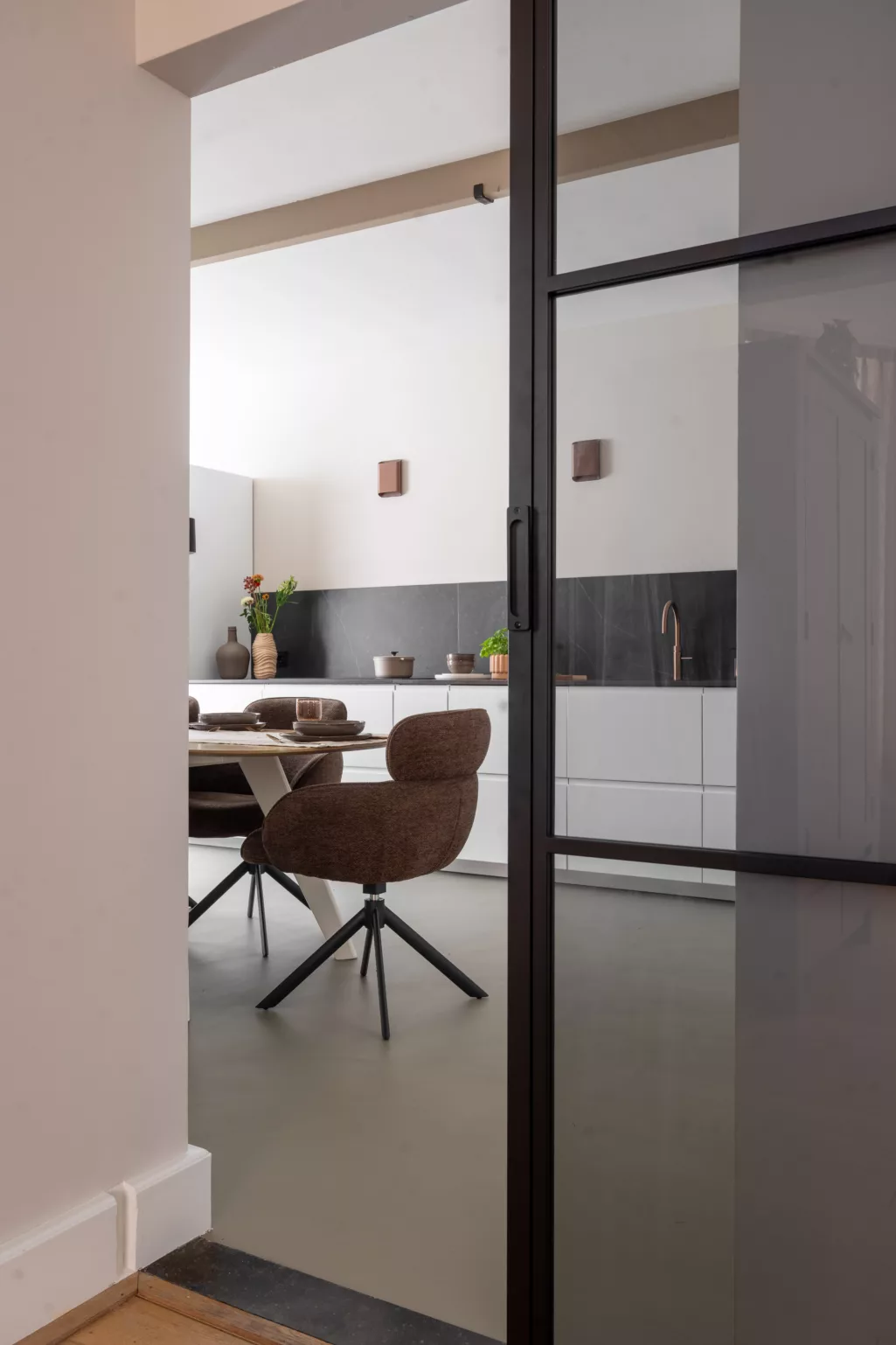 Doorkijk woonkamer, keuken, stalen schuifdeur rookglas, rechte keuken met witte onderkasten, Kelya dekton donker blad met bruine en grijze aderen, koperen wandlampen, beige wanden, grijze gietvloer