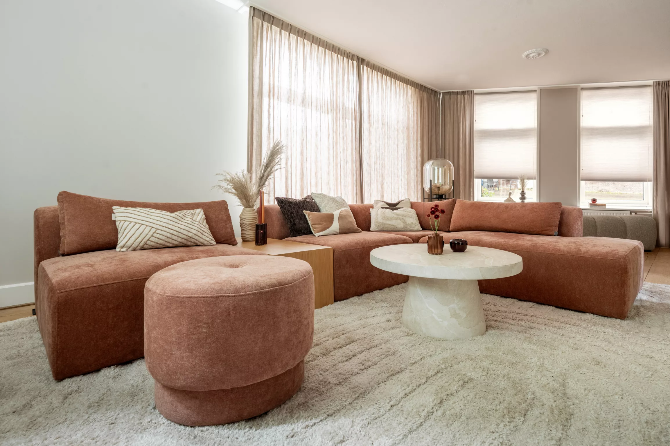 Grote woonkamer, terrakleurige hoekbank, elementenbank, tussentafel in licht eikenhout, bank staat volledig op vloerkleed, kleed beige met bogen, marmerlook tafel rond met stevige poot, ronde poef, beige in between gordijnen, plissé gordijnen in licht beige