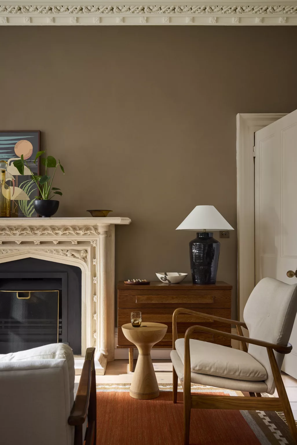 Zithoek bij openhaard met grote schouw, roestbruin kleed, klei kleurige wand, beige stoel met houten frame