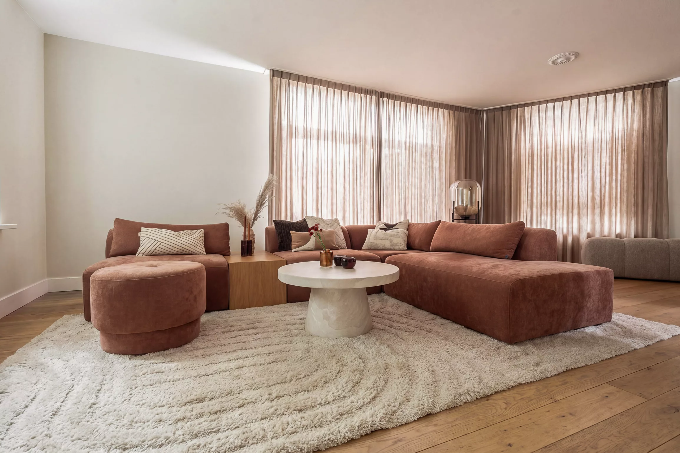 Ruime zithoek, roestbruine bank met bijpassende kussens en ronde poef, beige wanden en gordijnen.